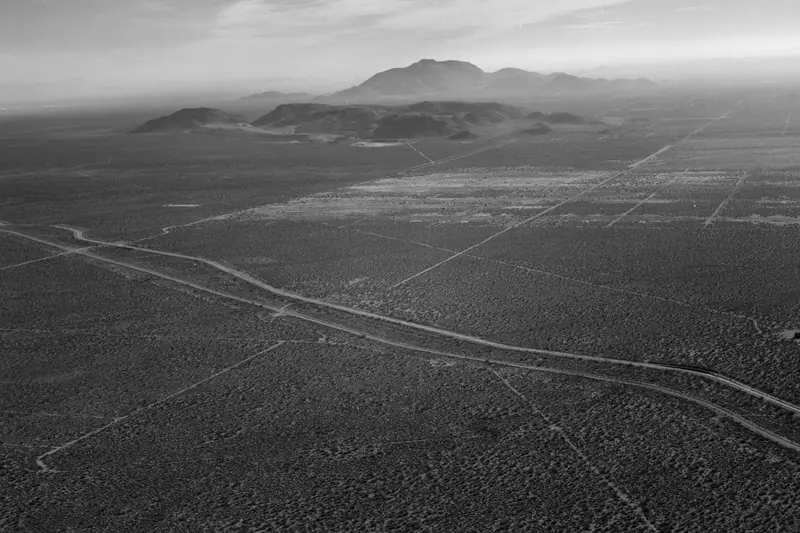 Black-and-white aerial view of two aqueduct lines running side by side across open desert scrub toward distant hills