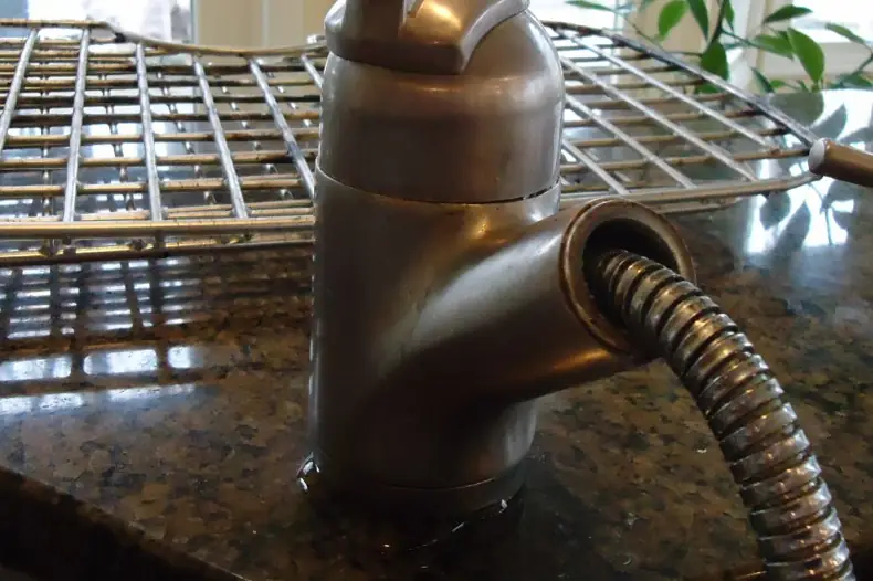 The base of a brushed bronze kitchen faucet on a granite counter, with a flexible metal supply hose coming out of its side