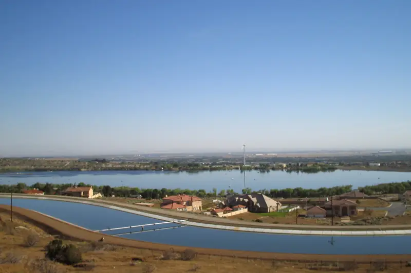 The California Aqueduct canal curving past Lake Palmdale, with houses on the strip of land between the canal and the lake under a clear sky