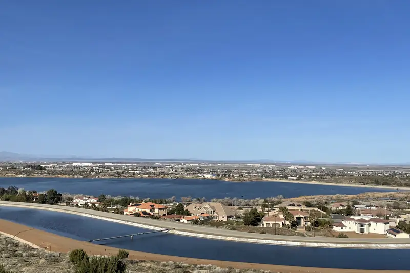 Lake Palmdale seen from a hillside, with the curving California Aqueduct canal and a row of houses in the foreground and the city spread across the flat valley beyond