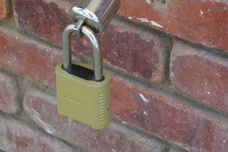 A hose spigot mounted on an outside brick wall, its handle locked with a small padlock