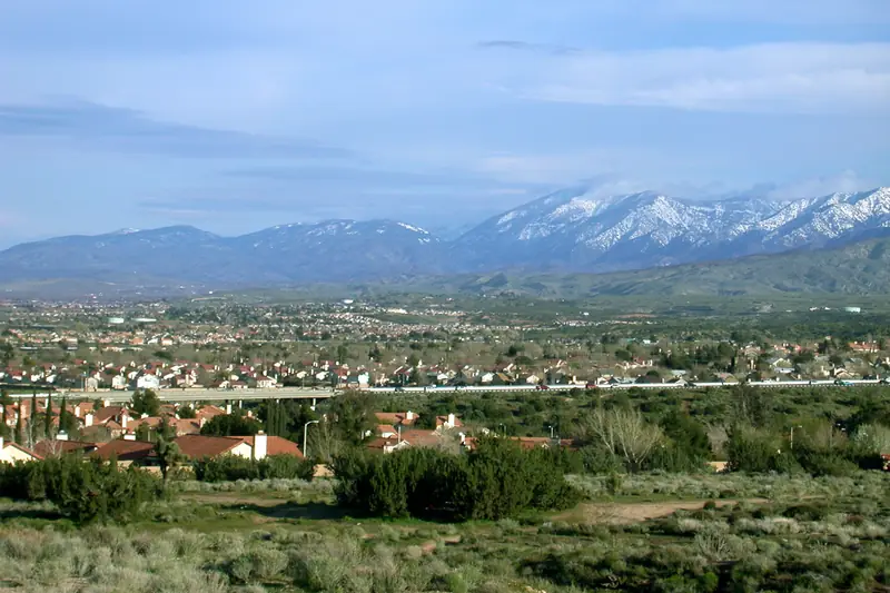 Rooftops of a Palmdale neighborhood spread across the valley floor, with snow-capped San Gabriel Mountains behind under a blue sky